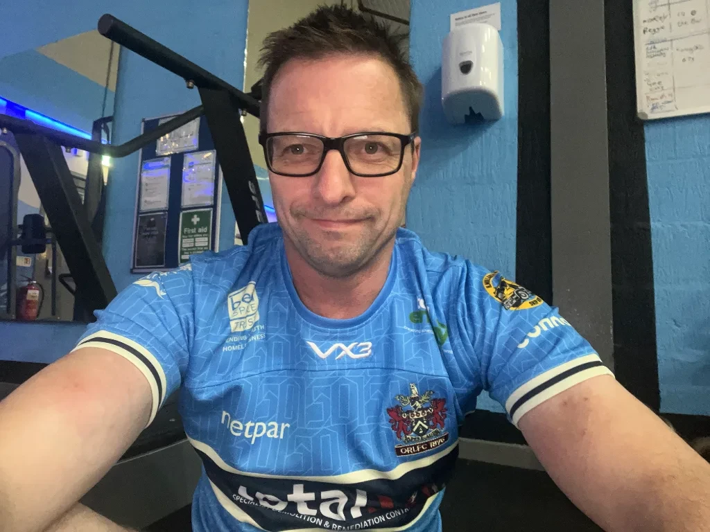 Man wearing glasses and blue rugby jersey with "netpar" branding takes a selfie in a gym with exercise equipment visible behind him.