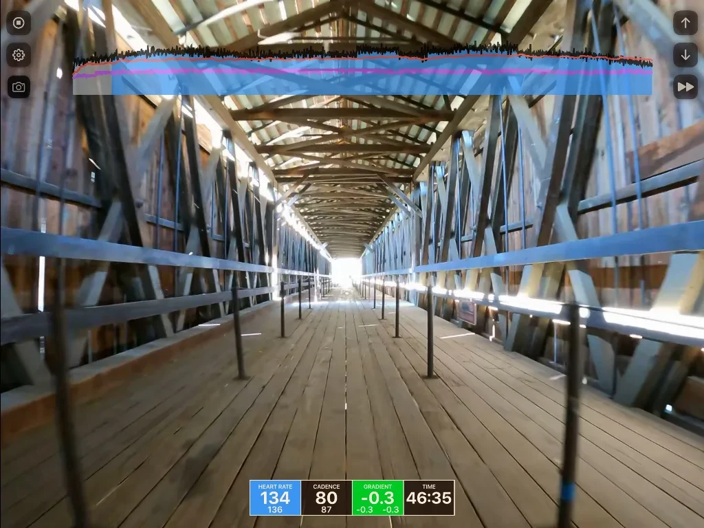 Long wooden covered bridge with metal railings stretching toward bright light, displaying heart rate 134, cadence 80, gradient -0.3, and time 46:35.