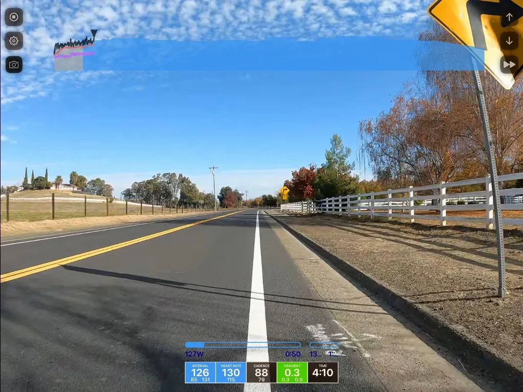 Straight rural road with white fencing on right, autumn trees, clear blue sky, dashboard metrics displaying speed, heart rate, and cadence data at bottom.