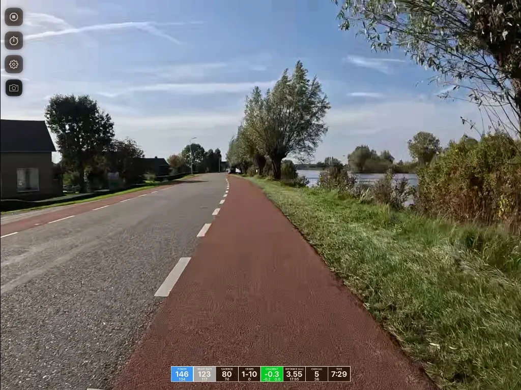 A red bike path runs alongside a gray road with a white center line, bordered by green grass and trees, with water visible on the right and a house on the left.