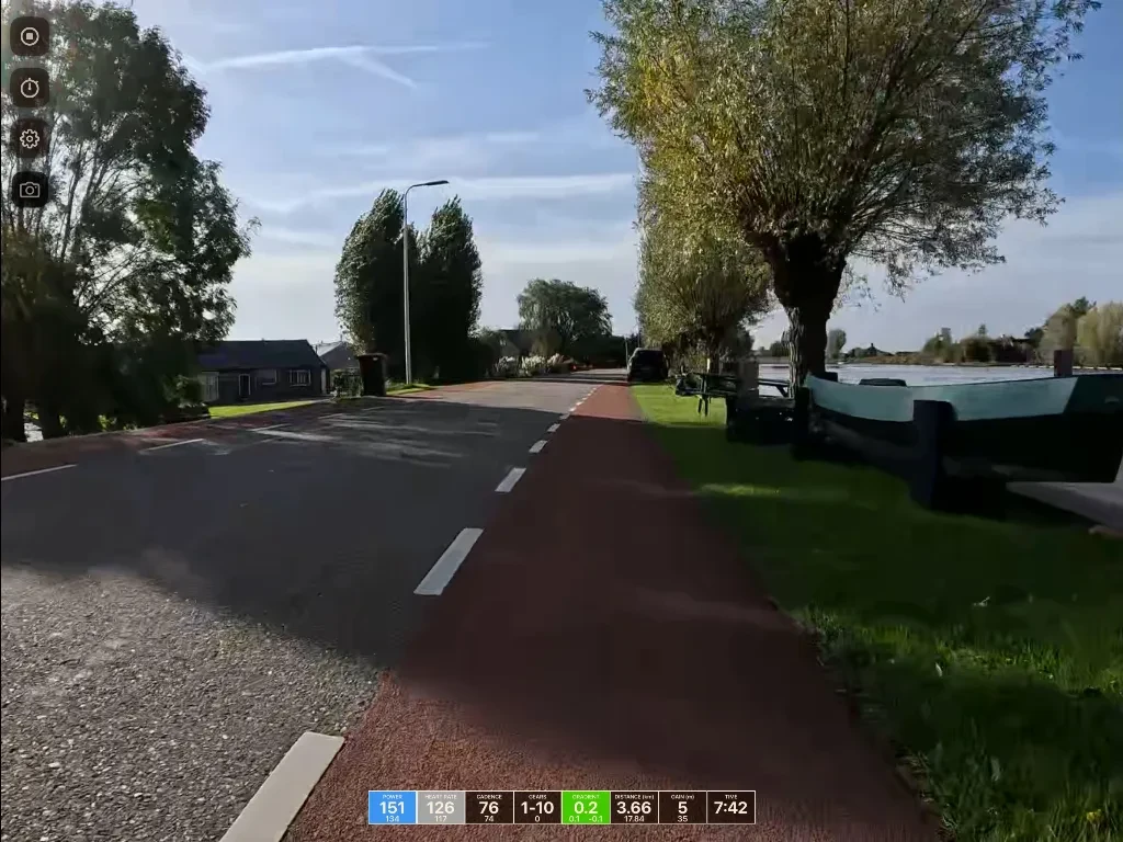 A rural Dutch road lined with tall trees, featuring a red bike path on the right, moored boats in a canal, and a cottage on the left under blue sky.