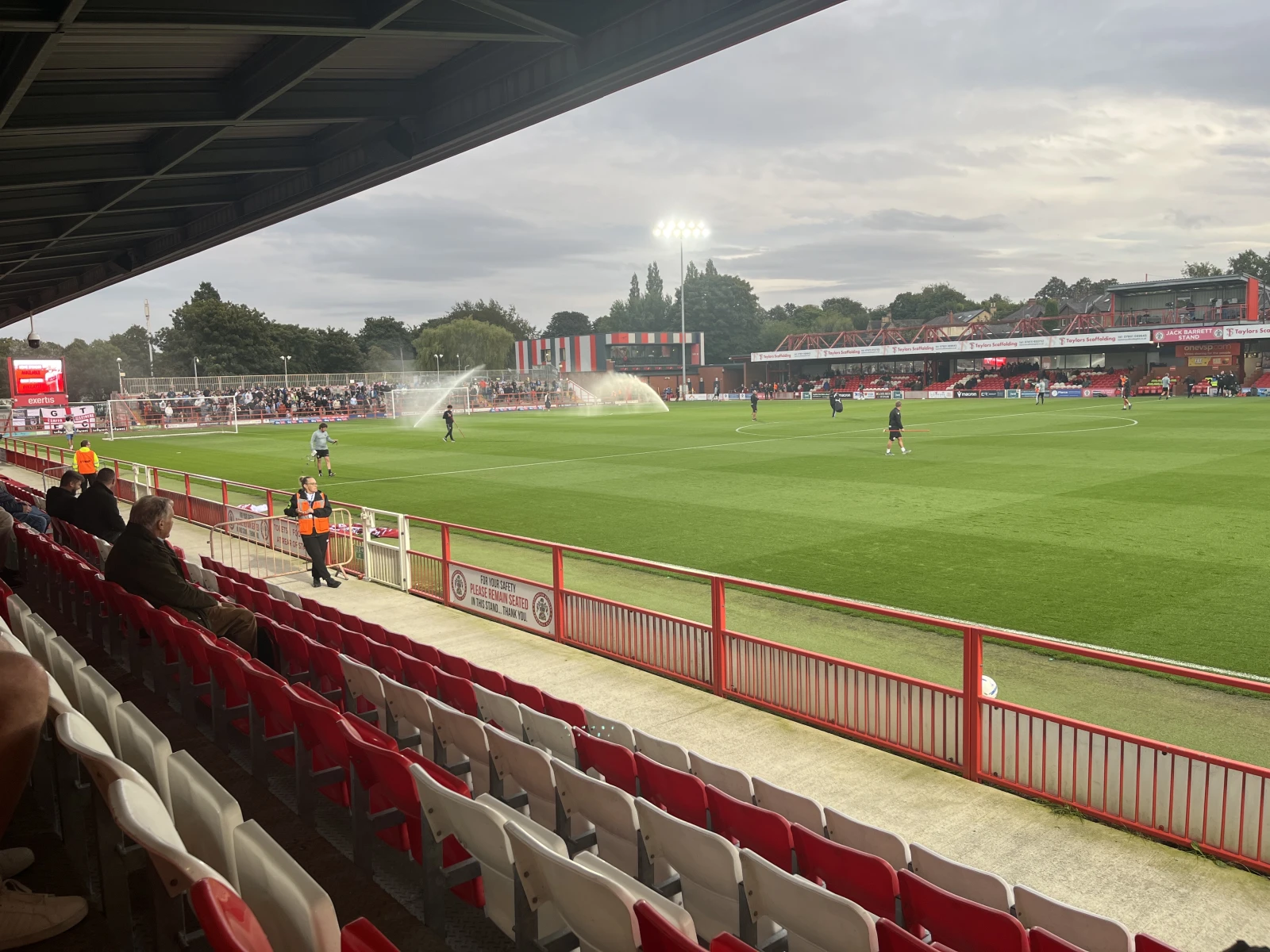 Red and white stadium seats in foreground, pitch with players and sprinkler, covered stand above, spectators in stands, trees and cloudy sky beyond.