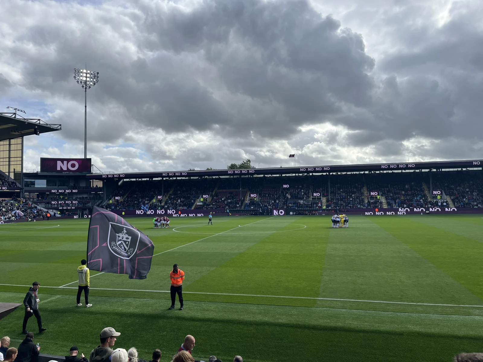 Football stadium with "NO" repeated on advertising boards, club flag on pitch, dramatic cloudy sky above, floodlight visible.