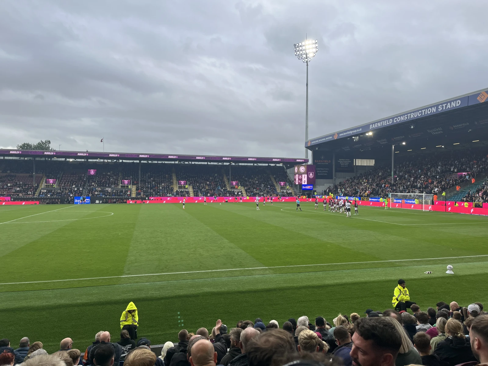 Barnfield Construction Stand at a football stadium during a match, with spectators in foreground and players on the pitch below overcast skies.
