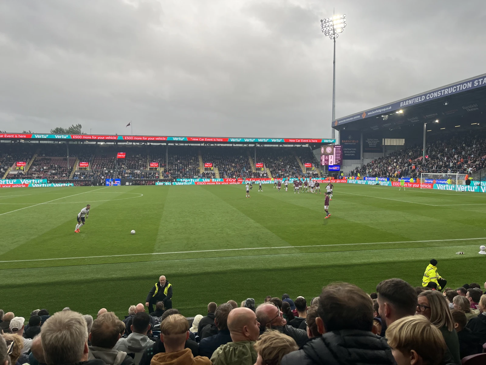 Football match in progress in front of the Barnfield Construction Stand with spectators in foreground, players on pitch, and advertising boards displaying Vertu branding.