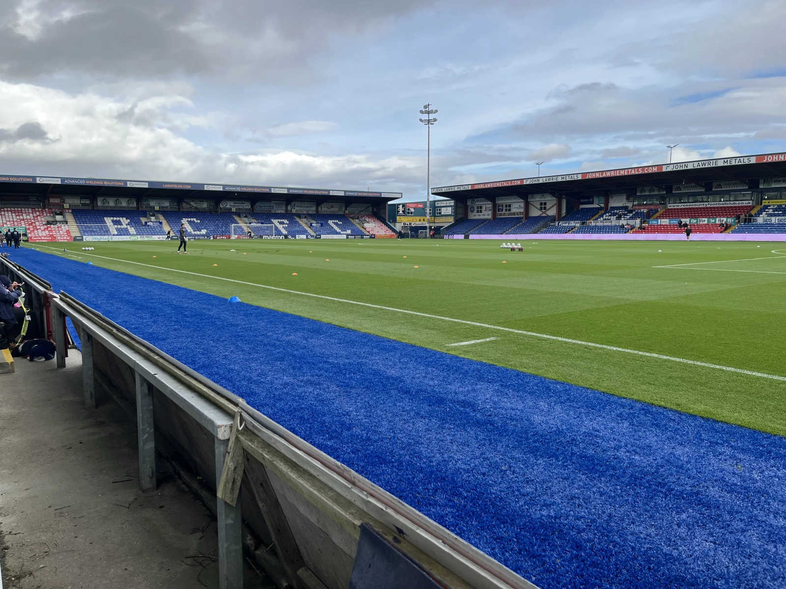 Blue pitch-side barrier in foreground, football stadium with red and blue seating, advertising boards reading "JOHN LAWEII METALS," empty grass pitch, tall floodlights, cloudy sky.