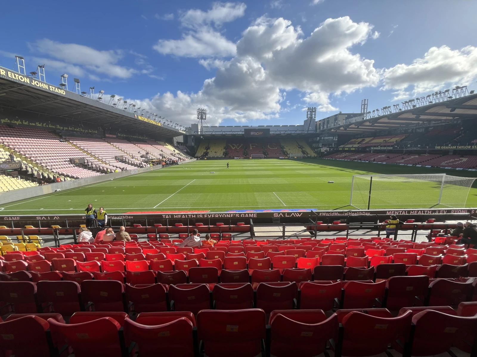 Red and yellow stadium seats in foreground, empty pitch beyond, "Belion John Stand" visible on upper left, advertising boards reading "Mental Health is Everyone's Game.