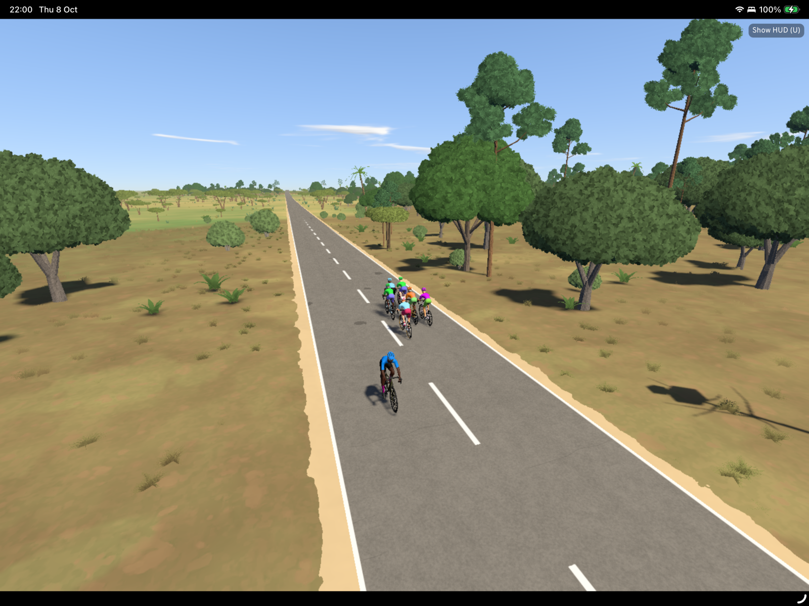 A straight asphalt road with white center lines stretches through a sparse savanna landscape dotted with acacia trees, with several cyclists riding on the road.